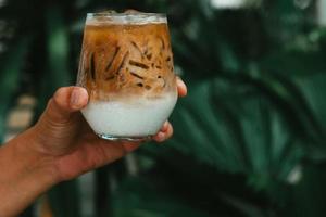 sostenga a mano café con leche helado en un vaso con crema en el fondo. fondo de bebida fría de verano foto