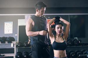 Personal fitness trainer coach helping asian woman lifting dumbbell top a head at inside gym club. photo