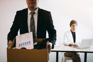 Front view with the male employee standing holding office supplies in the paper box going to submit a resignation letter while a female employee is working behind. photo