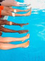 piernas de cuatro amigos de diferentes razas sentados junto a la piscina en un día soleado. fiesta. verano. concepto de vacaciones, amistad internacional y deporte. foto