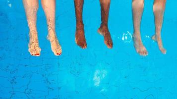 piernas de cuatro amigos de diferentes razas sentados junto a la piscina en un día soleado. fiesta. verano. concepto de vacaciones, amistad internacional y deporte. foto