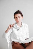 Woman with glasses and dressed in a white shirt is sitting on a chair with a book in her hands on a white background. Copy, empty space photo