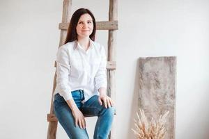 hermosa mujer vestida con una camisa blanca y jeans azules posando cerca de una escalera de madera sobre un fondo blanco. copiar, espacio vacío para texto foto
