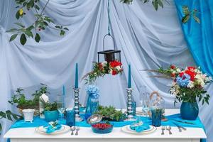 White banquet table covered with blue cloth, served with plates, cutlery, glasses, candles and flower arrangements. Copy, empty space for text photo