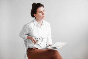 Beautiful woman with glasses and dressed in a white shirt is sitting on a chair with a book in her hands on a white background. Copy, empty space photo