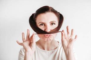 Portrait of a beautiful woman who makes a mustache out of pigtails and a funny face wearing a T-shirt on a white background. Copy, empty space photo