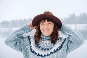 Portrait of a smiling woman in a brown hat and sweater on a background of snow-covered forest and lake photo