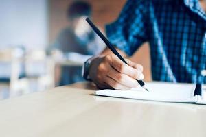 primer plano de la mano de un estudiante adolescente escribiendo en un examen en papel con un lápiz en el aula.