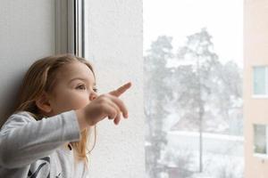 A little girl looks out the window at the flying snow and the winter snow-covered town photo