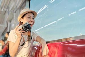 Beautiful Asian female tourist sits in a red seat, traveling by train, taking snapshot photo, transporting in suburb view, enjoy passenger lifestyle by railway, happy journey vacation. photo