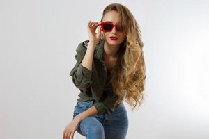 Stylish woman in red glasses looking at camera at studio isolated on white background. Copy space. Trendy style of beautiful luxury model with red lips makeup. Long wavy shiny blonde hair photo