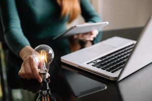 business hand holding lightbulb with using laptop computer and money stack in office. idea saving energy and accounting finance concept photo