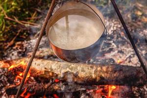 Cast iron pot cooks over open fire in a campsite in forest in sunny summer day. Camping tourism relax trip active lifestyle adventure travel picnic concept. photo