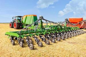 Agricultural seeders attached to tractors stand on yellow stubble in a wide field against a blue cloudy sky on a summer day. photo