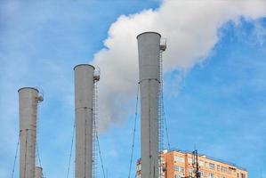 Three chimneys of a thermal power plant emitting steam under a clear blue sky against the backdrop of the top of a residential building. photo