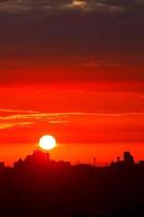 A bright red sun disk rises in the early morning over the silhouettes of city houses. Vertical image. photo