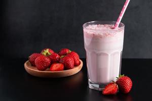 Milkshake with strawberries on black background. Summer drink in a glass with a paper straw.