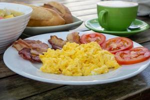 Close Up Food Photo of Breakfast with Scrambled Eggs, Bacon Strips, Tomatoes, Baguette, Cappuccino and Fruit Bowl on a Wooden Table