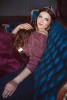 portrait of beautiful young pretty cute woman princess  in long purple queen's dress and crown, with long hair and make up sitting on the blue velvet sofa indoor in minimalistic interior