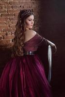 portrait of beautiful young pretty cute woman princess in long purple queen's dress and crown, with long hair and make up indoor in loft interior near leather chair photo