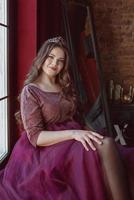 portrait of beautiful young pretty cute woman princess in long purple queen's dress and crown, with long hair and make up indoor in loft interior sitting by the window photo