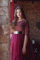 portrait of beautiful young pretty cute woman princess in long purple queen's dress and crown, with long hair and make up indoor in minimalistic loft interior standing by the window photo