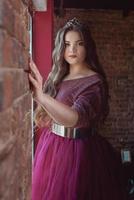 portrait of beautiful young pretty cute woman princess in long purple queen's dress and crown, with long hair and make up indoor in minimalistic loft interior standing by the window photo