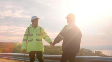 people engineer are shaking hand in a field wearing a protective helmet on her head over electrical turbines background . video