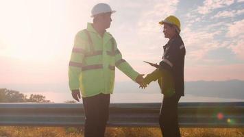 people engineer are shaking hand in a field wearing a protective helmet on her head over electrical turbines background . video