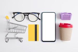 Online shopping flat lay creative composition. Knolling with phone mockup, supermarket trolley, credit card, coffee cup and glasses photo