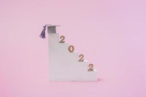 Simple concept of education on a pink background. Graduation 2022. Podium steps with graduation cap and wooden numbers closeup. photo