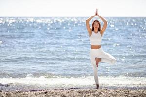 mujer joven haciendo yoga en la playa con ropa blanca foto