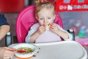 Mother hand feeding infant girl with spoon in high baby chair in kitchen at home photo