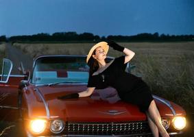A beautiful girl poses on the hood of a red convertible Caddy, evening, sunset.