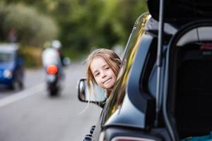 Little funny girl grimaces, leaning out of the car window. Family travel by car in Europe. photo