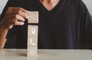 hand holding wooden cube block icon letter telephone location and address on table.Business communication and social network concept. photo