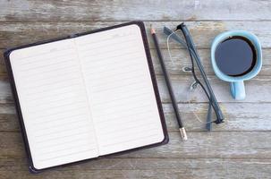 Blank paper notebook,glasses,pencil and cup of coffee on brown wooden table background.