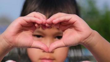 close-up van een klein Aziatisch meisje dat naar de camera kijkt en een hartvorm maakt met haar handen. hartsymbool van liefde. video