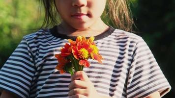 een schattig klein meisje ruikt de bloemen van een boeket bloemen in haar hand op een zonnige zomerdag. gelukkig kind genieten van de natuur buitenshuis. geur zintuiglijk leren van bloem. video