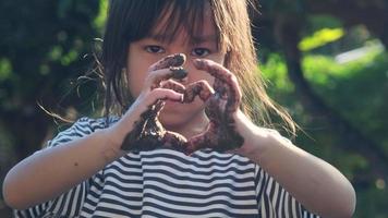 schattig klein meisje met modderige handen maakt een hartvorm met haar handen tegen de achtergrond van de natuur. concept van natuur en liefde symbool. video