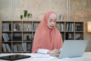 Beautiful businesswoman of Asian ethnicity works on e-commerce using laptop, internet communication in small business office. Attractive person, traditionally Islam dresses by wearing hijab. photo