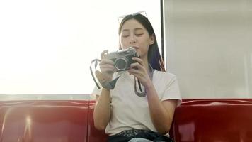 Beautiful Asian female tourist with camera sits in red seat, traveling by sky train, taking snapshot photo when transporting in urban view, city passenger lifestyle by railway, happy journey vacation. video