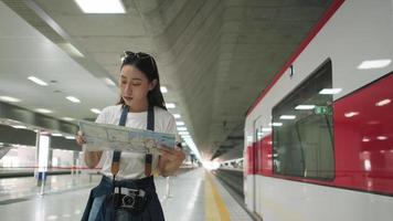 uma bela turista asiática com óculos e câmera, procurando no mapa de localização do viajante, procurando e encontrando destino na estação de trem pública da tailândia, transporte ferroviário, estilo de vida casual. video