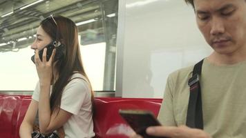 hermosa turista asiática con cámara sentada en un asiento rojo, viajando en tren aéreo, hablando por teléfono móvil cuando se transporta en vista urbana, estilo de vida de pasajeros de la ciudad en tren, feliz viaje de vacaciones. video