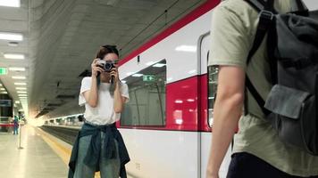 Young beautiful Asian female tourist taking photo with film camera with male friend, smile and enjoyment at train station platform, happy travel lifestyle by subway transport vacation trip. video
