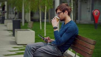 Outdoor scene of handsome young man browsing smartphone social network app while drink coffee. video