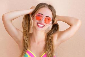 cerca de una chica feliz con gafas de sol rosas aisladas. vacaciones de verano y fin de semana divertido. concepto de verano. mujer joven sonriente en traje de baño de moda. enfoque selectivo. estilo de traje de verano de playa. foto