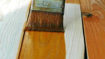 o pintor aplica tecnicamente a mancha de madeira na placa de madeira texturizada recém-aplainada com um pincel no fundo da oficina. trabalho de artesão de madeira profissional lento e preciso. fechar-se. vista lateral video