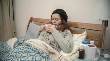 mujer hispana acostada sola en su cama bien envuelta, enferma de gripe, bebiendo una taza de té foto