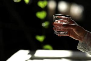 manos de mujer aisladas, sosteniendo un vaso de agua sobre un fondo oscuro con hojas verdes. los rayos del sol caen sobre el cristal. mañana saludable foto